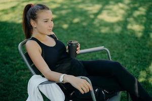 Außenaufnahme einer nachdenklichen dunkelhaarigen schönen Frau in Sportkleidung trinkt Kaffee zum Mitnehmen verbringt Freizeit in Open-Air-Posen auf einem Stuhl im Garten genießt gutes Wetter atmet frische Luft foto