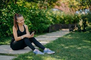 Zufriedene sportliche Frau in Sportkleidung lächelt fröhlich, surft auf dem Handy, während sie Musik über drahtlose Kopfhörer hört, macht eine Pause, nachdem das Training am sonnigen Morgen die App zur Aktivitätsverfolgung verwendet foto