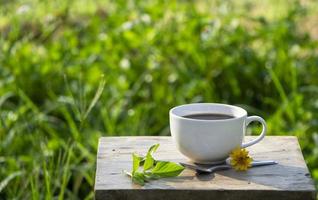 Hochwinkelansicht, eine weiße Tasse schwarzen Americano-Kaffee auf einem rustikalen Holztisch mit grünem Naturhintergrund und Morgensonne. Konzept für Erfrischungsgetränke foto