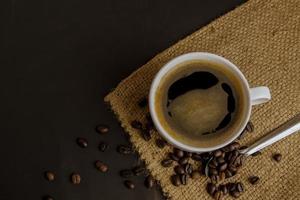 Draufsicht auf eine weiße Keramiktasse mit heißem Kaffee aus schwarzem Americano auf Sackleinen und einem dunklen Hintergrund mit einem Haufen gerösteter Kaffeebohnen foto