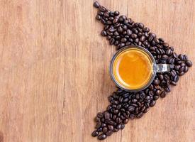 Blick von oben auf eine Tasse heißen Espressokaffees mit einer schönen Crema auf einem Holztisch, umgeben von einem Haufen braun gerösteter Kaffeebohnen in Pfeilform foto