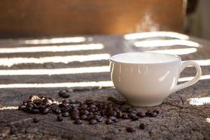 Weiße Kaffeetasse aus Keramik auf einem rustikalen Holztisch mit Kaffeebohnen unter der Morgensonne durch den Zaun. foto