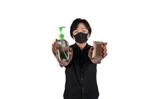 asiatischer mann hält und zeigt reinigungsalkoholgel und seife auf seiner hand und trägt eine schwarze maske auf klarem weißem hintergrund. foto