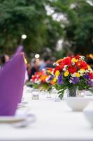 Rote, gelbe, violette und weiße Blumen stehen auf dem langen, weißen Deckentisch und sind bereit für ein luxuriöses Abendessen im Gartenfeld. foto