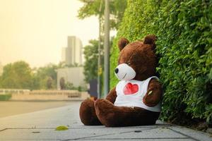 Dunkelbraune Bärenpuppe, die in einsamer Stimmung auf dem Fußweg neben der Straße sitzt. foto