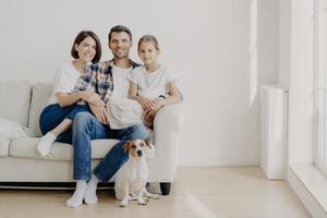 horizontale Ansicht von drei freundlichen Familienmitgliedern und einem kleinen Rassehund posieren vor der Kamera, um unvergessliche Fotos zu machen. tochter verbringt freizeit mit mutter und vater, schönen tag zu hause