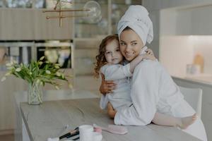 Foto der jungen Mutter und Tochter, die sich umarmen und ihre Liebe ausdrücken, in einer modernen Wohnung posieren, weiche weiße Bademäntel tragen, sich kosmetischen Eingriffen unterziehen, gesunde Haut haben. Zeit für Schönheit