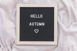 herbstlicher Hintergrund. schwarzes buchstabenbrett mit textphrase hallo herbst auf weißem strickpullover liegend. Ansicht von oben, flach liegend. Thanksgiving-Banner. Hygge-Stimmungskonzept für kaltes Wetter foto