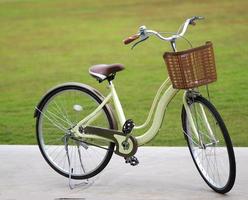 Das gelbe Fahrrad hat einen Korb aus braunem Kunststoff, der auf Betonboden geparkt ist, verschwommen vom Naturhintergrund foto