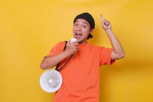 asiatischer junger mann im lässigen stil mit megaphon und nach oben zeigendem finger isoliert auf gelbem hintergrund. foto