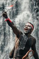 geist von papua mann, der traditionelle kleidung des dani-stammes, rot-weißes stirnband und armreif trägt, hält kleine indonesien-flagge und feiert den indonesischen unabhängigkeitstag vor wasserfallhintergrund. foto