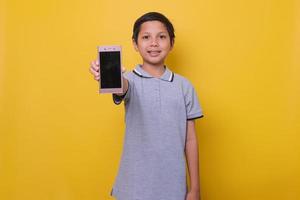 asiatischer junge im lässigen stil lächelt und zeigt den bildschirm des smartphones isoliert auf gelbem hintergrund. kopieren sie platz und verspotten sie für online-lernen und kindermodekonzept. foto