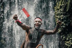 papua-mann des dani-stammes lächelt und feiert den indonesischen unabhängigkeitstag vor wasserfallhintergrund. foto