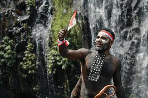 geist von papua mann, der traditionelle kleidung des dani-stammes, rot-weißes stirnband und armreif trägt, hält kleine indonesien-flagge und feiert den indonesischen unabhängigkeitstag vor wasserfallhintergrund. foto