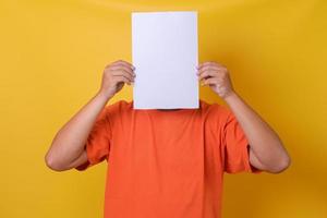 junger Mann in orangefarbenem T-Shirt, der sein Gesicht unter leerem weißem Papier versteckt, isolierter gelber Hintergrund. foto