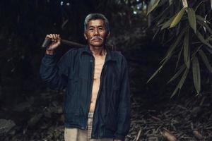 Portait des asiatischen Bauern zeigt Daumen nach oben und trägt Hacke vor dunklem Vintage-Hintergrund. foto