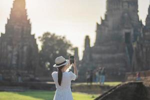glückliche touristische frau im weißen kleid, das foto mit mobilem smartphone macht, während des besuchs im wat chaiwatthanaram-tempel im historischen park ayutthaya, sommer-, solo-, asien- und thailand-reisekonzept