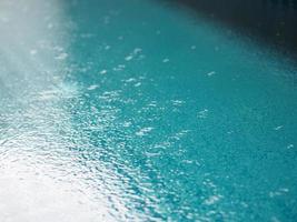 Regentropfen, die blaues Wasser im Poolhintergrund fallen lassen, kräuseln sich auf der Oberflächenstruktur, glitzernde Bokeh-Zusammenfassung foto