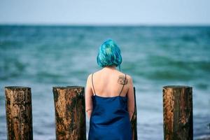 Schöne blauhaarige Frau in langem dunkelblauem Kleid, die am Sandstrand steht und den Meereshorizont betrachtet foto