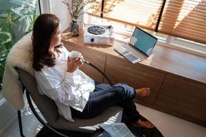 Fröhliche, lässige, schöne asiatische Frau, die an einem Laptop arbeitet, während sie auf einem Stuhl im Wohnzimmer bei ihr zu Hause sitzt. foto