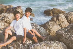 homosexuelles porträt junges asiatisches paar sitzt umarmung zusammen auf felsen oder stein am strand im sommer, asien homosexueller tourismus für freizeit und entspannen mit glücklichem urlaub auf meer, lgbt-rechtskonzept. foto