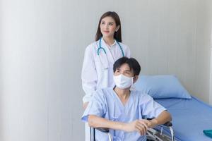 asiatische ärztin, die mit einem männlichen patienten spricht, der eine gesichtsmaske über sein gesundheitssymptom trägt, während er im krankenhaus im rollstuhl sitzt. foto