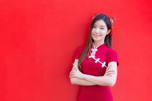 schöne asiatische frau trägt ein rotes cheongsam und einen gekreuzten arm, während sie in die kamera schaut und glücklich mit dem roten hintergrund lächelt. foto