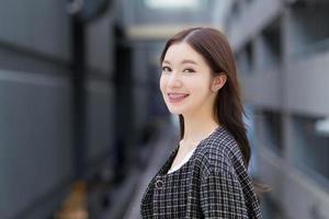 Porträt einer professionellen jungen asiatischen Geschäftsfrau in einem schwarzen Mustermantel mit Zahnspangen, die draußen in der Stadt mit einem Bürogebäude im Hintergrund steht und lächelt. foto
