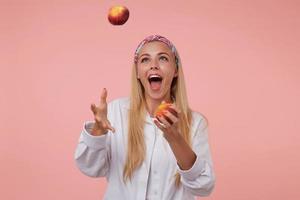 fröhliche hübsche dame mit langen blonden haaren, die im studio mit pfirsichen jongliert, spaß auf rosa hintergrund hat, farbiges stirnband und weißes hemd trägt foto