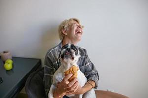 Attraktives, fröhliches blondes Mädchen, das sich mit ihrem schwarz-weißen Hund amüsiert, das Haustier versucht, Croissant zu probieren und freut sich auf die Kamera, sitzt im Esszimmer foto