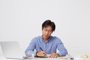 Nachdenklich fokussierter asiatischer junger Geschäftsmann mit Brille, der am Tisch mit Laptop arbeitet und Plan in Notizbuch über weißem Hintergrund schreibt foto