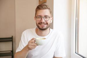 Nahaufnahme eines schönen Mannes mit einer Tasse in der Hand, der eine Brille und Kopfhörer trägt, aufrichtig in die Kamera lächelt und über dem Innenraum posiert foto