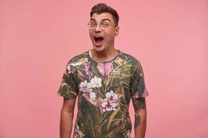 Schöner junger Mann mit Tätowierungen in lässigem farbigem T-Shirt, der mit den Händen nach unten über rosafarbenem Hintergrund steht, aufgeregt und überrascht aussieht, die Stirn zusammenzieht und den Mund weit öffnet foto