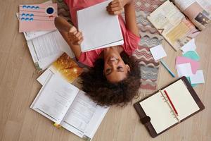 Draufsicht einer attraktiven jungen lockigen Frau mit dunkler Haut, die auf dem Teppich liegt, während sie Notizen liest, sich mit vielen Büchern auf Prüfungen vorbereitet und ein rosa T-Shirt trägt foto