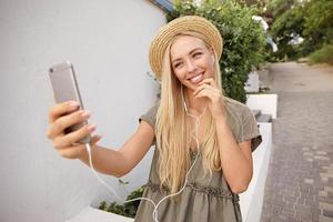 junge glückliche blonde frau, die musik hört, während sie selfie mit ihrem handy macht, lässiges leinenkleid und strohhut trägt, fröhlich und erfreut aussieht foto