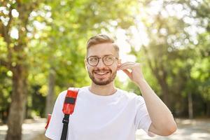 Porträt eines gutaussehenden, fröhlichen Mannes, der wegschaut und breit lächelt, Musik über Kopfhörer hört und an einem warmen und sonnigen Tag über dem grünen Park posiert foto