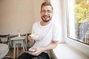 junger attraktiver typ in weißem t-shirt mit bart, der mit sanftem lächeln zur kamera schaut, neben fenster sitzt und kaffee trinkt, sms schreiben will foto