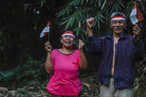 indonesisches älteres ehepaar, das gemeinsam den indonesischen unabhängigkeitstag im dorf feiert. alter mann und frau mit rot-weißem stirnband und kleiner indonesischer flagge. foto