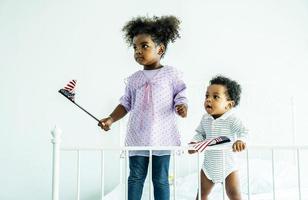 glücklicher süßer aktiver kleiner afroamerikanischer kinderjunge und mädchen, die amerikanische flagge auf dem bett halten foto