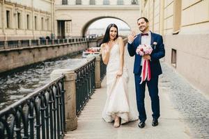 Aufgeregtes Ehepaar zeigt Ringe an den Fingern, geht auf der Brücke, posiert vor der Kamera, um unvergessliche Fotos zu machen. brünette frau im weißen kleid und gutaussehender mann hält brautblumenstrauß-show-ringe