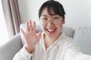 junge asiatische frau, die smartphone für online-videokonferenzanrufe mit freunden verwendet, die mit der hand winken und eine hallo-geste machen foto