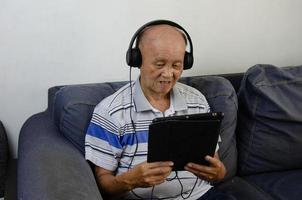 älterer mann, der ein tablet auf dem sofa hält und musik mit kopfhörern und sozialen medien hört. foto