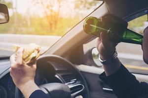 Mann fährt Auto, während er Hamburger isst und Bier trinkt foto