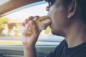 Mann isst gefährlich Hotdog und kaltes Getränk beim Autofahren foto