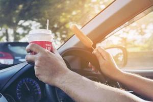 Mann, der Auto fährt, während er Kaffee und Hotdog hält, schlechtes und gefährliches Verhalten foto