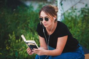 Foto eines fröhlich lächelnden, süßen jungen Studentenmädchens, das im Freien eine Sonnenbrille trägt und mit dem Handy plaudert, um Musik mit Kopfhörern zu hören.