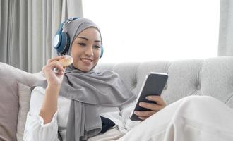 attraktive junge asiatische muslimische frau, die hijab und kopfhörer trägt, filme auf dem smartphone anschaut und donuts auf dem sofa isst. foto