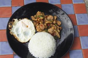 basilikum, hähnchen, spiegelei, thailändisches essen der hausstil ist nicht gehoben in einer schwarzen keramikplatte, die auf einem karierten betontisch steht. servierfertig und versandfertig foto