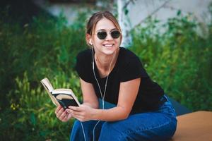 Foto eines fröhlich lächelnden, süßen jungen Studentenmädchens, das im Freien eine Sonnenbrille trägt und mit dem Handy plaudert, um Musik mit Kopfhörern zu hören.