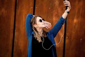 Foto eines fröhlich lächelnden, süßen jungen Studentenmädchens, das im Freien eine Sonnenbrille trägt und mit dem Handy plaudert, um Musik mit Kopfhörern zu hören.